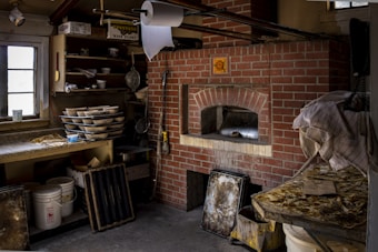 A rustic kitchen with a brick pizza oven is surrounded by various kitchen items. Stacks of baking trays and bowls are on shelving, and a large roll of paper towels hangs from the ceiling. The counter is cluttered with baking essentials and reflective metal containers. The overall setup conveys a functional, slightly cluttered baking workspace.