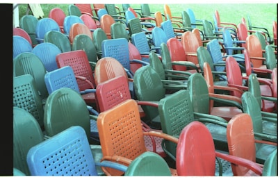 A large number of colorful plastic chairs are stacked haphazardly in an outdoor setting, with the main colors being green, red, blue, and orange.