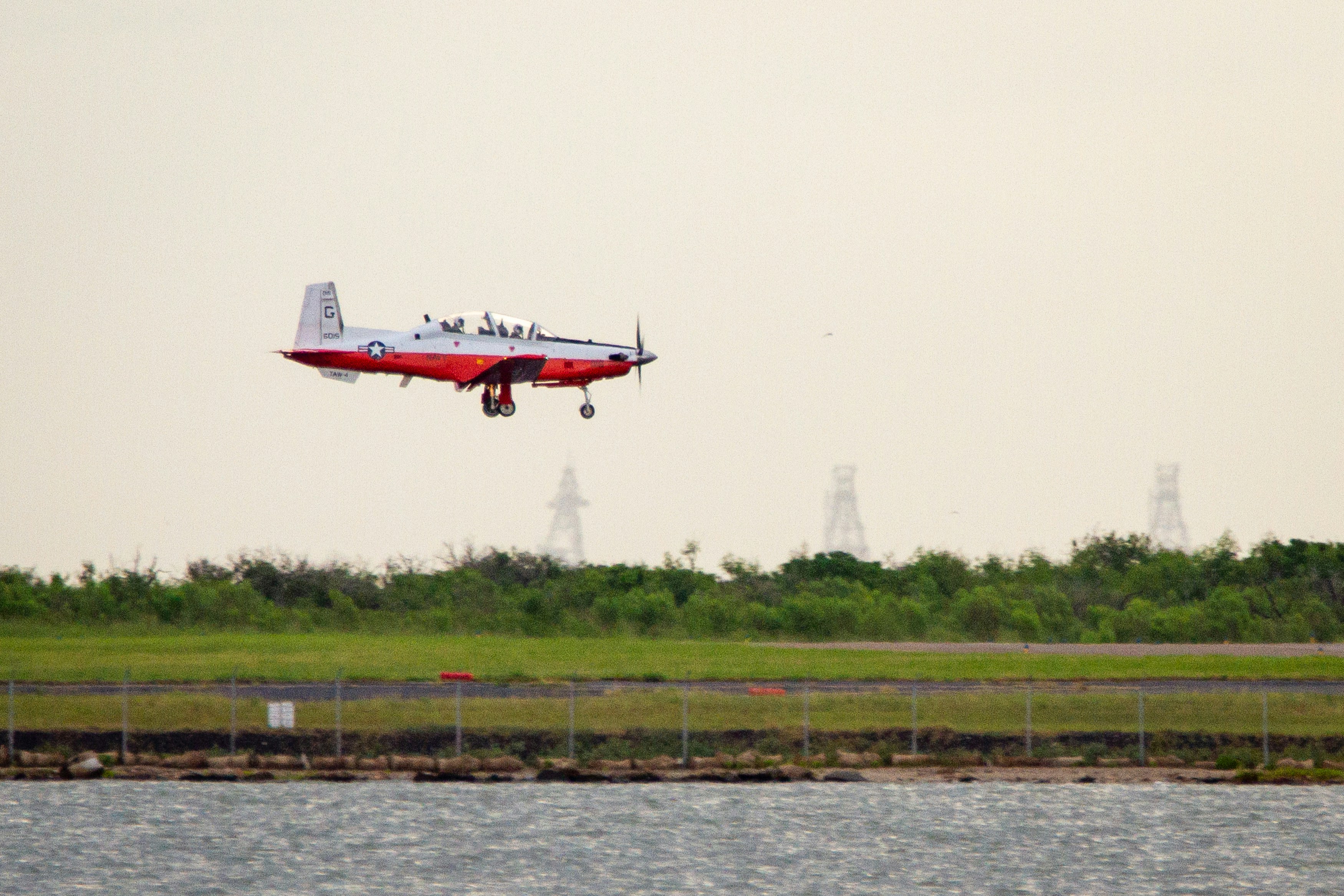 white and red airplane flying over the sea during daytime, A T-6B Texan 11 comes in for a landing at Naval Air Station Corpus Christi.