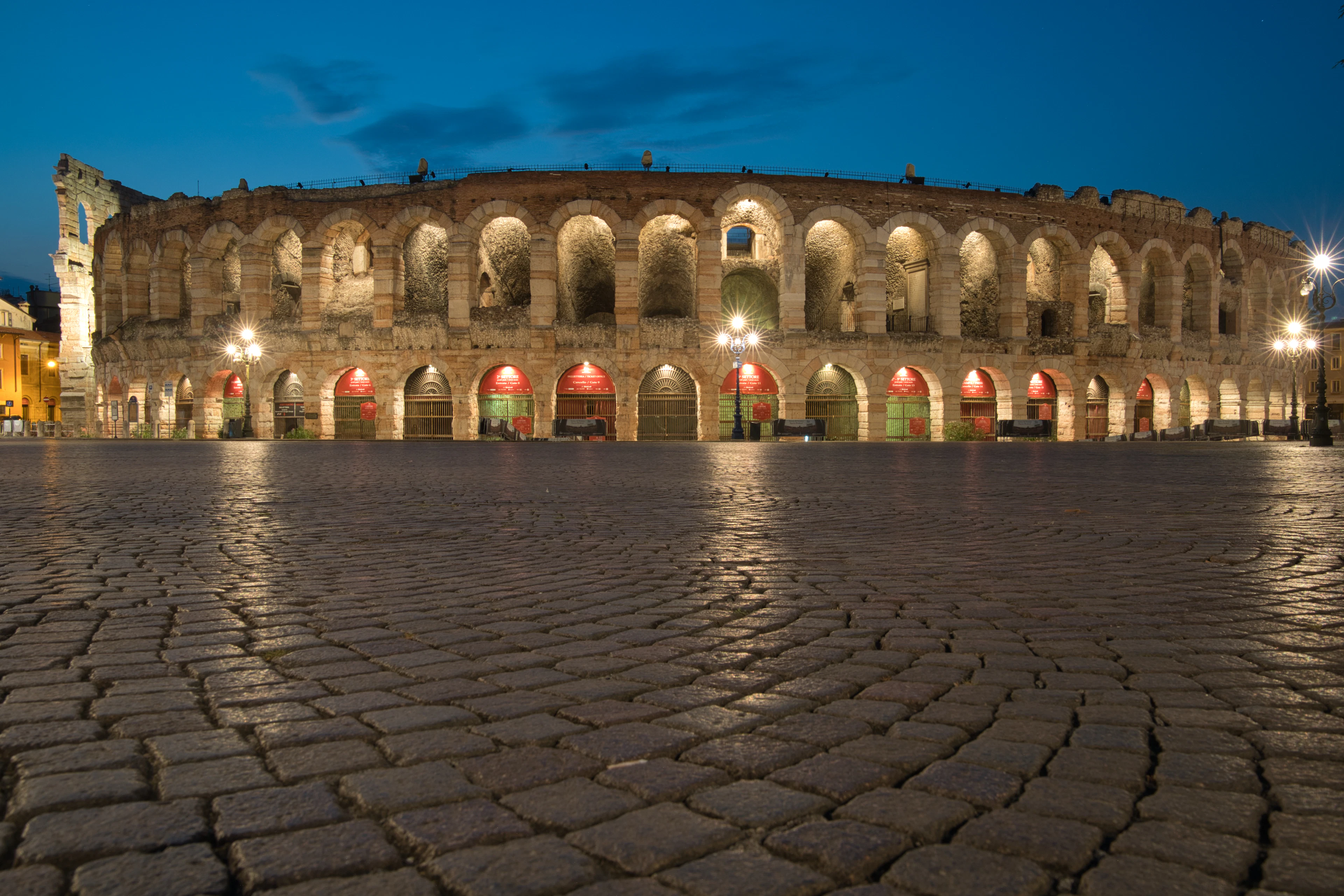 Verona Arena illuminated at dusk beside an empty cobblestone piazza with glowing lamps and red awnings.