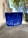 A vibrant blue paint streak is applied on what appears to be a transparent surface like glass. The background shows a blurred outdoor scene with trees and a building, which seems to be out of focus.