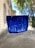 A vibrant blue paint streak is applied on what appears to be a transparent surface like glass. The background shows a blurred outdoor scene with trees and a building, which seems to be out of focus.