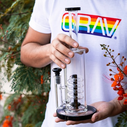 A person is holding a glass spiral percolator bong with a black base. They are wearing a white t-shirt with a colorful rainbow logo. In the background, there are green plants and orange flowers.