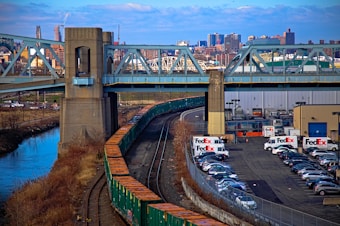 A bridge with a freight train passing underneath stretches across a river amidst an urban skyline. In the background, a FedEx facility occupies the center right, with multiple FedEx trucks and a parking lot. Several buildings and city infrastructure are visible in the distance under a partially cloudy sky.
