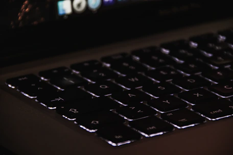 Close-up of a laptop keyboard illuminated by soft turquoise backlight in a dim room.