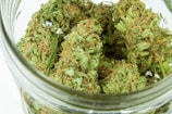 A close-up view of dried cannabis buds stored in a glass jar. The buds are dense with vibrant green leaves and orange hairs, showing visible crystals and a sticky appearance.