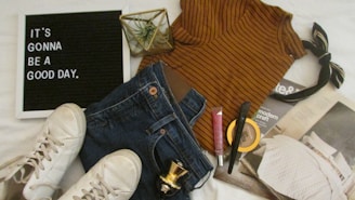 A casual outfit arrangement includes a striped brown top, blue jeans, white sneakers, a black and beige hair bow, and various cosmetic items such as lip gloss, foundation, and mascara. A decorative plant and a magazine are also present. A black letter board displays the inspiring message 'It's gonna be a good day.'