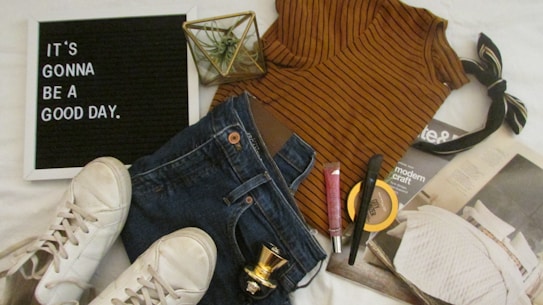 A casual outfit arrangement includes a striped brown top, blue jeans, white sneakers, a black and beige hair bow, and various cosmetic items such as lip gloss, foundation, and mascara. A decorative plant and a magazine are also present. A black letter board displays the inspiring message 'It's gonna be a good day.'