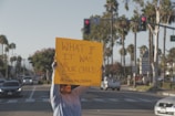 A person stands on a street holding a bright yellow sign that reads 'WHAT IF IT WAS YOUR CHILD? #SavetheChildren'. The background shows a typical urban setting with palm trees, cars, and traffic signals.