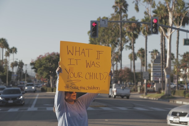 A person stands on a street holding a bright yellow sign that reads 'WHAT IF IT WAS YOUR CHILD? #SavetheChildren'. The background shows a typical urban setting with palm trees, cars, and traffic signals.