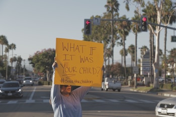 A person stands on a street holding a bright yellow sign that reads 'WHAT IF IT WAS YOUR CHILD? #SavetheChildren'. The background shows a typical urban setting with palm trees, cars, and traffic signals.