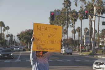 A person stands on a street holding a bright yellow sign that reads 'WHAT IF IT WAS YOUR CHILD? #SavetheChildren'. The background shows a typical urban setting with palm trees, cars, and traffic signals.