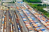 Aerial view of a large cargo terminal filled with colorful shipping containers organized in rows. Railway tracks are visible, some with train cars loaded with containers. The area is surrounded by industrial buildings and greenery.