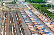 Aerial view of a large cargo terminal filled with colorful shipping containers organized in rows. Railway tracks are visible, some with train cars loaded with containers. The area is surrounded by industrial buildings and greenery.