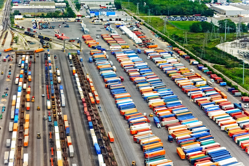 Aerial view of a large cargo terminal filled with colorful shipping containers organized in rows. Railway tracks are visible, some with train cars loaded with containers. The area is surrounded by industrial buildings and greenery.