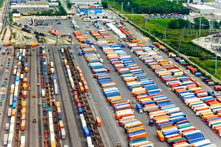 Aerial view of a large cargo terminal filled with colorful shipping containers organized in rows. Railway tracks are visible, some with train cars loaded with containers. The area is surrounded by industrial buildings and greenery.