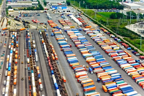 Aerial view of a large cargo terminal filled with colorful shipping containers organized in rows. Railway tracks are visible, some with train cars loaded with containers. The area is surrounded by industrial buildings and greenery.