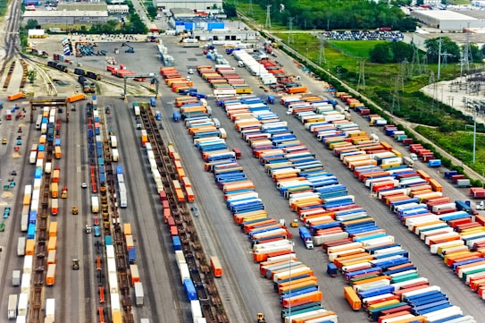 Aerial view of a large cargo terminal filled with colorful shipping containers organized in rows. Railway tracks are visible, some with train cars loaded with containers. The area is surrounded by industrial buildings and greenery.