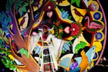 A person is dressed in colorful traditional attire, standing in front of a vibrant and decorative backdrop featuring various elements like birds, flowers, and geometric patterns. The artwork includes vivid motifs of nature such as trees, leaves, chameleons, and flowers, all contributing to a lively and festive atmosphere.