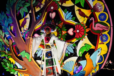 A person is dressed in colorful traditional attire, standing in front of a vibrant and decorative backdrop featuring various elements like birds, flowers, and geometric patterns. The artwork includes vivid motifs of nature such as trees, leaves, chameleons, and flowers, all contributing to a lively and festive atmosphere.