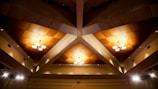 A geometric ceiling design with wooden panels arranged in a triangular pattern. The ceiling features several modern chandeliers, each with multiple round lights. The overall lighting creates a warm glow, and the edges of the ceiling are equipped with small spotlights.
