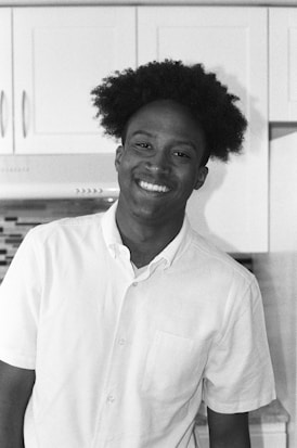 A person is smiling warmly in a kitchen setting, dressed in a white shirt. The background includes white cabinets and a countertop.