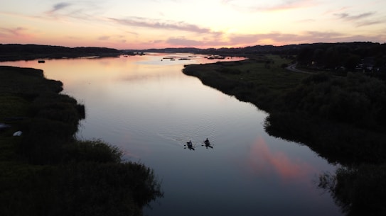 A serene river scene during sunset with two kayakers paddling along calm waters. The sky is painted with soft hues of pink, orange, and purple, reflecting beautifully on the river's surface. Lush greenery lines the riverbanks, fading into silhouettes against the colorful sky.