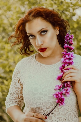 A person with red hair is wearing a white lace dress and holding a branch of pink flowers. The background is a blurred green foliage, giving a natural and serene setting.