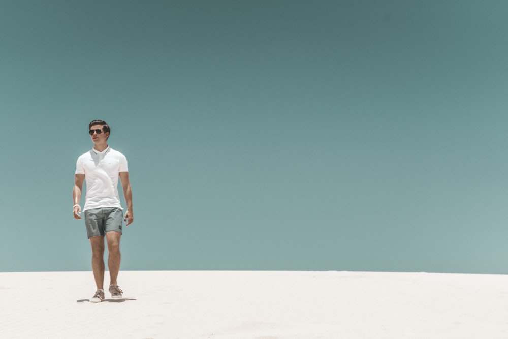man in white t-shirt and blue denim shorts standing on white sand during daytime