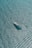 Aerial view of a large yacht gliding through a vast expanse of shimmering blue water. The sunlight reflects off the water, creating a sparkling effect across the surface. The yacht, with its sleek design, is positioned slightly off-center, moving diagonally across the frame.