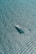 Aerial view of a large yacht gliding through a vast expanse of shimmering blue water. The sunlight reflects off the water, creating a sparkling effect across the surface. The yacht, with its sleek design, is positioned slightly off-center, moving diagonally across the frame.