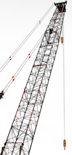 A tall construction crane with a lattice structure, stretching diagonally across the frame. The crane is equipped with several pulleys and cables, showcasing its industrial design.