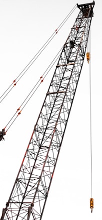 A tall construction crane with a lattice structure, stretching diagonally across the frame. The crane is equipped with several pulleys and cables, showcasing its industrial design.
