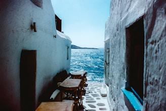 A narrow cobblestone alley leads down to a clear blue sea. Whitewashed buildings frame the pathway, with wooden tables and chairs positioned along the side. The bright sunlight casts distinct shadows and the vibrant color of the sea contrasts against the stark walls.