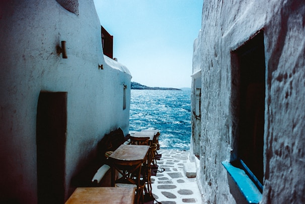 A narrow cobblestone alley leads down to a clear blue sea. Whitewashed buildings frame the pathway, with wooden tables and chairs positioned along the side. The bright sunlight casts distinct shadows and the vibrant color of the sea contrasts against the stark walls.