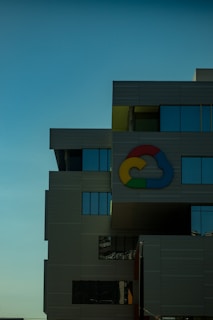 A modern building with a gridlike facade and numerous rectangular windows reflecting the sky. A colorful cloud-like logo is prominently displayed on the building, against a clear blue sky in the background.