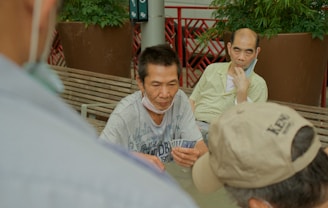 Several individuals gather outdoors, sitting on benches and engaging in a game of cards. The focal point is on a man holding a fan of playing cards, with others observing or participating. Potted plants and a red railing are visible in the background, contributing to the setting's casual and social atmosphere.
