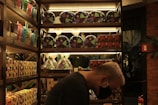 A person wearing a black mask is standing in front of well-organized shelves stocked with various packaged goods, including gift baskets wrapped in cellophane and colorful containers. The setting appears to be a store, with warm lighting highlighting the products neatly displayed on wooden shelves. In the background, there is a glimpse of greenery through a window and a fire extinguisher visible on the right side.