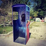 A creatively painted phone booth stands outdoors on a paved area. The booth features artwork of a red dog and abstract blue and white patterns. The environment includes green trees and piles of materials in the background.