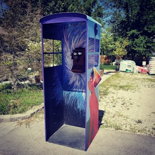 A creatively painted phone booth stands outdoors on a paved area. The booth features artwork of a red dog and abstract blue and white patterns. The environment includes green trees and piles of materials in the background.