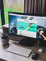 A modern home office setup featuring a large monitor displaying a webpage, with a wireless keyboard and mouse on a wooden desk. A small potted succulent is positioned to the left of the monitor. Natural light filters in through a window with closed curtains on the right.