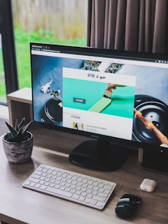 A modern home office setup featuring a large monitor displaying a webpage, with a wireless keyboard and mouse on a wooden desk. A small potted succulent is positioned to the left of the monitor. Natural light filters in through a window with closed curtains on the right.