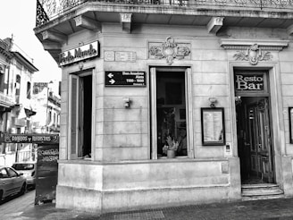 A black and white photo depicts the corner section of an old building with intricate architectural details. There is a 'Resto Bar' entrance, potted plants visible inside through a window, and signage with directions to streets named Don Anselmo and Aieta. The facade has ornate sculptures above the windows and a decorative iron balcony railing on the upper floor.
