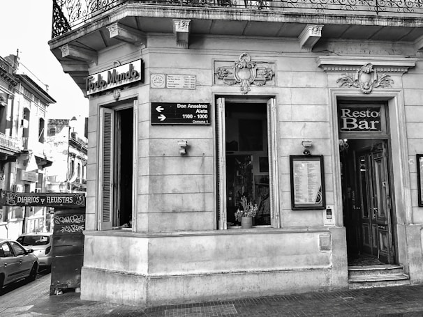 A black and white photo depicts the corner section of an old building with intricate architectural details. There is a 'Resto Bar' entrance, potted plants visible inside through a window, and signage with directions to streets named Don Anselmo and Aieta. The facade has ornate sculptures above the windows and a decorative iron balcony railing on the upper floor.