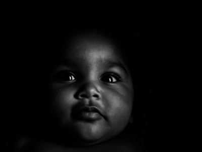 A baby is captured in a low light setting, creating a dramatic contrast with its facial features highlighted against a dark background.