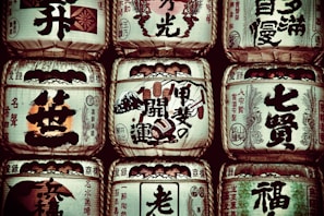Several traditional sake barrels are stacked in a symmetrical arrangement. Each barrel is adorned with Japanese calligraphy and symbols, highlighting intricate designs and various insignias representative of sake breweries.