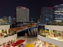 A nighttime cityscape featuring a river surrounded by tall, illuminated buildings. In the foreground, there are two levels of outdoor seating areas with red umbrellas and tables. The reflections of the buildings' lights dance on the water, creating a vibrant and lively atmosphere.