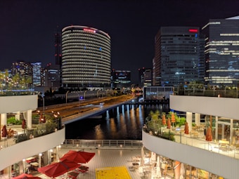 A nighttime cityscape featuring a river surrounded by tall, illuminated buildings. In the foreground, there are two levels of outdoor seating areas with red umbrellas and tables. The reflections of the buildings' lights dance on the water, creating a vibrant and lively atmosphere.