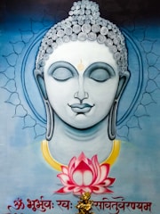 A serene and artistic depiction of a spiritual figure with closed eyes and a peaceful expression. The detailed hair is adorned with spiral patterns, and a subtle golden aura encircles the head, adding to the ethereal quality. Below, a vibrant pink lotus flower is depicted, surrounded by calm blue tones. Intricate designs and inscriptions in a script are present at the bottom of the image.