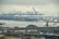 A large cargo port featuring numerous cranes and containers, with a prominent Yang Ming Line cargo ship near the docks. Several smaller boats are visible in the water, and an elevated highway runs across the foreground. Overcast skies create a muted atmosphere.